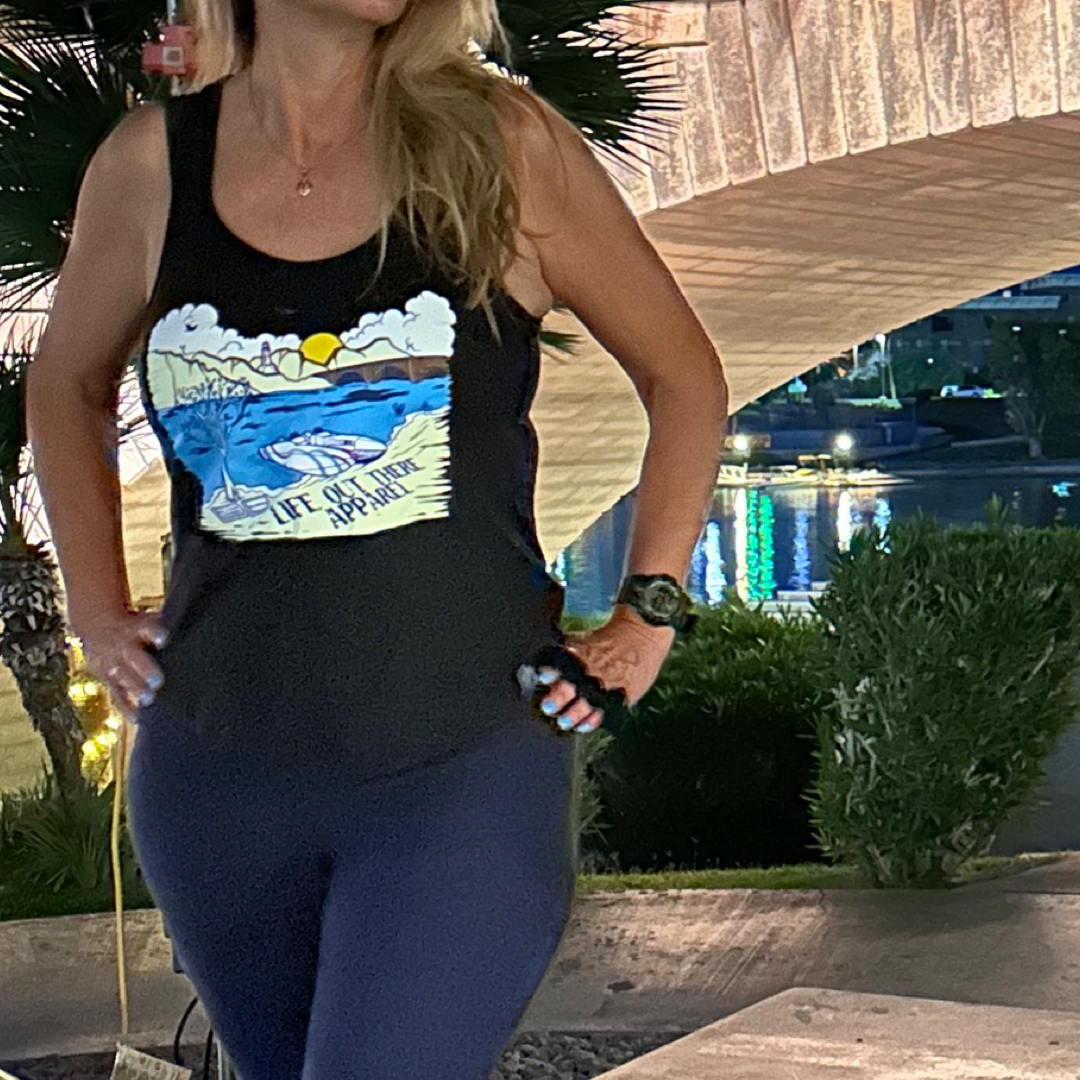 Under the bright lights near London Bridge, a person with long hair stands confidently, hands on hips, showcasing the stunning scenery of water and trees in the background. They are wearing The Havasu Women's Tank Top in black by Life Out There Apparel, featuring an eye-catching graphic design.