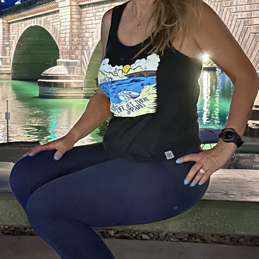 A person wearing The Havasu Women's Tank Top in black from Life Out There Apparel pairs it with dark leggings while sitting by Lake Havasu at night. Behind them, a bridge with illuminated arches creates shimmering reflections on the water, as they enjoy the tranquil scene with their face turned away.
