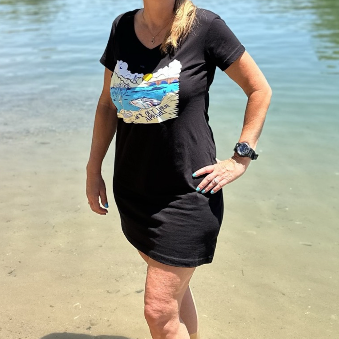 Standing on the edge of Lake Havasu, a person wears The Havasu Women's Cover Up in black from Life Out There Apparel. The vibrant design features a sunset, waves, and a cloud. Their arms rest by their sides as they gaze at the calm water under the clear sky.