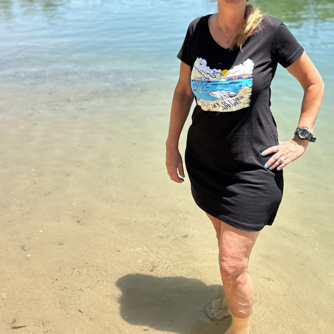 A woman stands in shallow water at the beach near London Bridge, wearing The Havasu Women's Cover Up in black from Life Out There Apparel. The vibrant graphic design on the cover-up complements the sunny day, creating a clear reflection on the water.