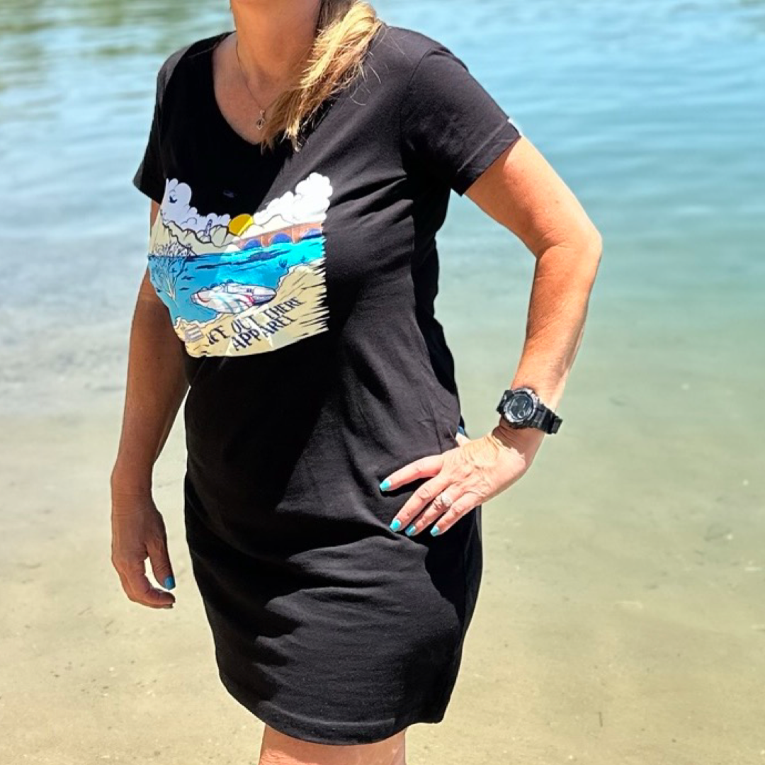 A person wearing The Havasu Women's Cover Up in black, featuring a vibrant beach scene print from Life Out There Apparel, stands by Lake Havasu. The individual sports light nail polish and a black watch. In the background, the calm water with gentle ripples beautifully encapsulates the serene atmosphere of this unique location.