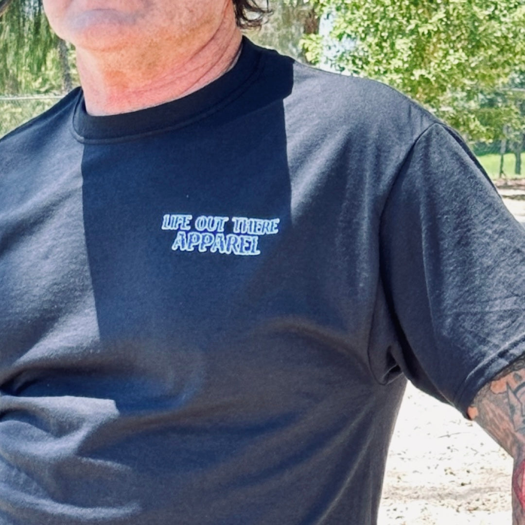 A person wearing the Unisex The Havasu T Shirt in black from Life Out There Apparel stands outdoors, capturing those summer lake vibes. Sunlight creates shadows on the shirt, while green trees and a fence form a scenic backdrop that echoes Lake Havasu's serene charm.