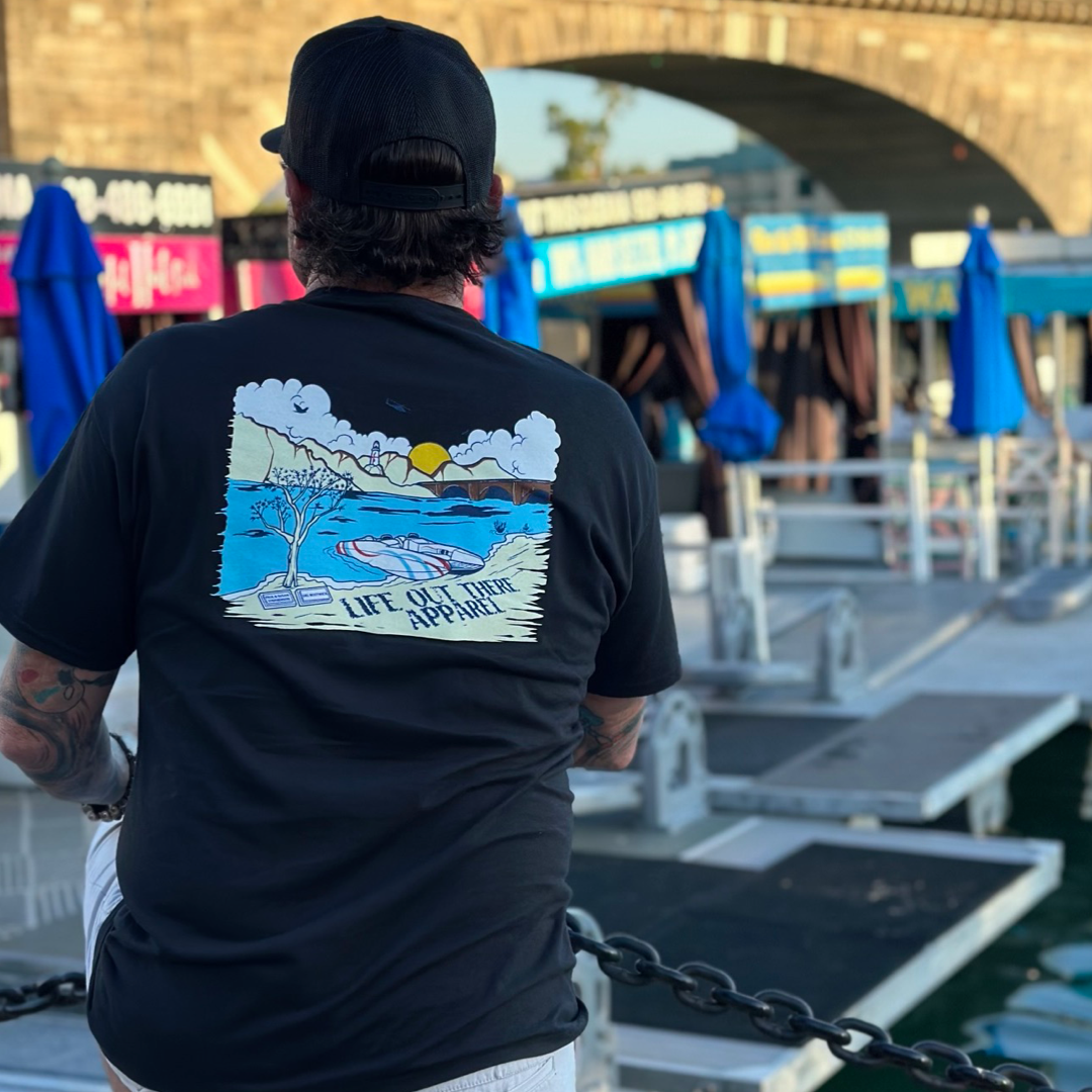 A person wearing the Unisex The Havasu T Shirt - Black by Life Out There Apparel and a black cap stands by a chain fence, taking in the summer lake vibes of boat docks and blue umbrellas. An arched stone bridge adds charm to the scene, evoking memories of serene days at Lake Havasu.