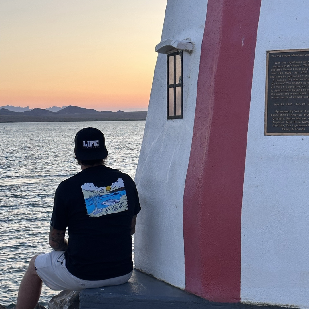 Wearing the Unisex The Havasu T Shirt in black from Life Out There Apparel, a person sits by the red and white striped lighthouse, soaking in the summer lake vibes while watching the sunset over a serene Lake Havasu with distant mountains on the horizon.