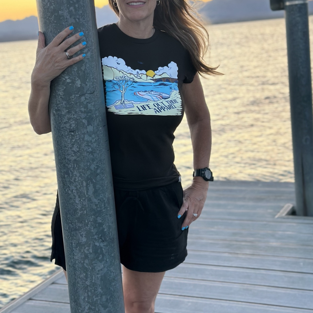 A woman stands on a wooden dock by the water at sunset, exuding summer vibes. She wears Life Out There Apparel's The Havasu Women's T Shirt in black, paired with black shorts. Her left hand rests on a vertical pole, while Lake Havasu's calm waters and distant hills provide the perfect backdrop.