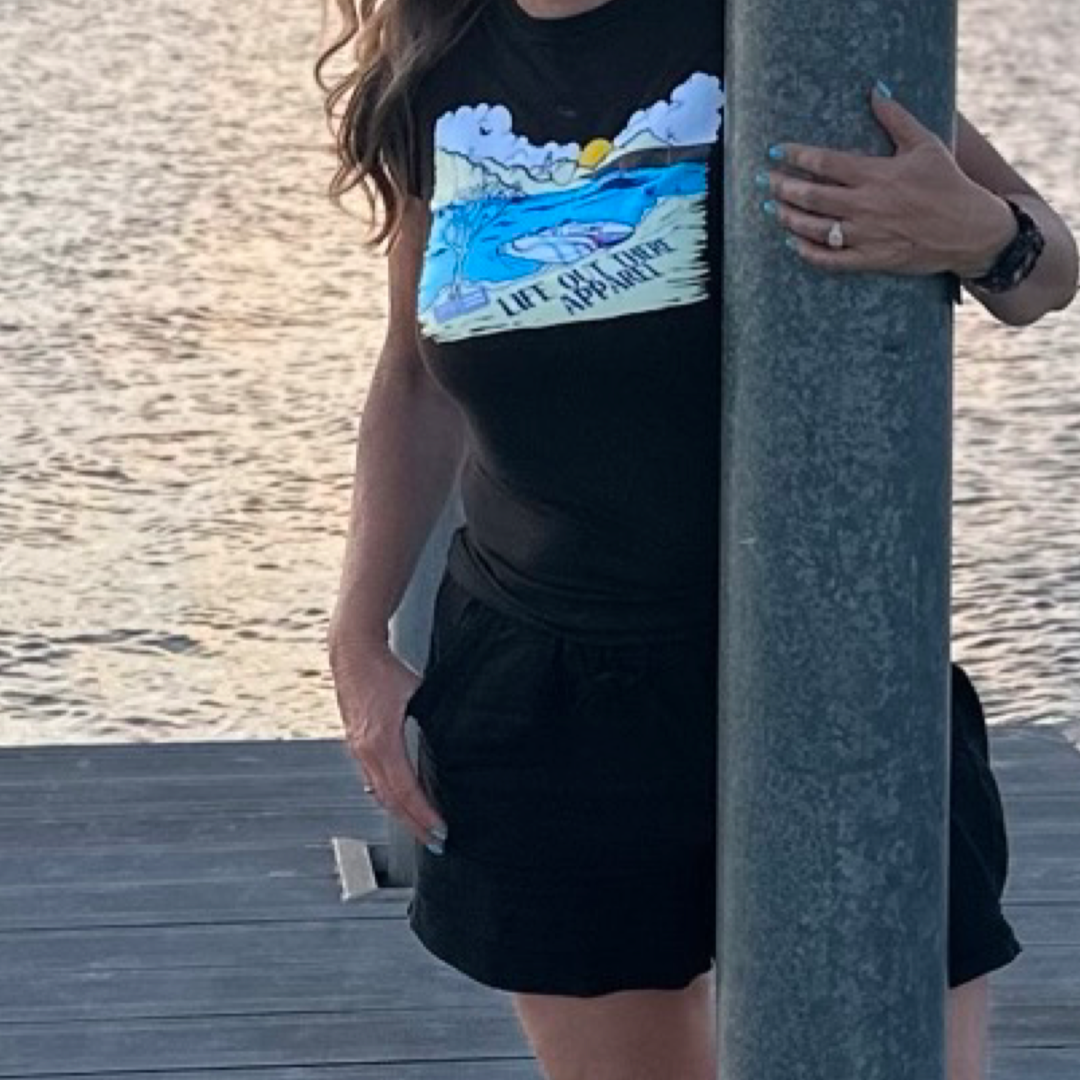 Wearing *The Havasu Women's T Shirt - Black* by Life Out There Apparel, a person leans against a wooden post on a dock, with the shimmering Lake Havasu stretching out behind them. Their other hand is casually tucked in their pocket, their shorts swaying gently in the breeze and exuding those perfect summer vibes.