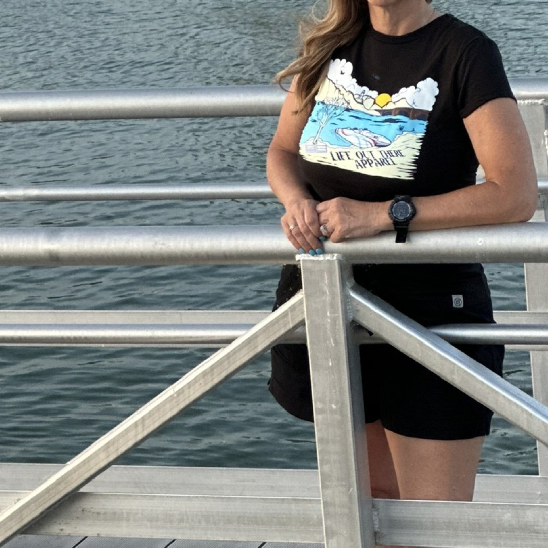 A person wearing *The Havasu Women's T Shirt - Black* from Life Out There Apparel stands on a dock by Lake Havasu, leaning on a metal railing. The shirt design showcases a cloud, sun, and waves, perfectly capturing the summer vibes.