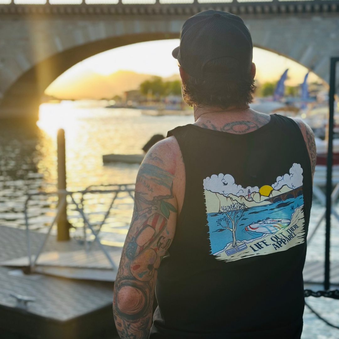 A person adorned with tattoos and a cap stands by the waterfront, gazing at the sunset over Lake Havasu. They're wearing The Havasu Men's Tank Top in black from Life Out There Apparel, which features a vibrant design of a beach scene with the phrase "Life OutThere Apparel," perfectly encapsulating those summer lake vibes. In the background, a bridge and boats can be seen.