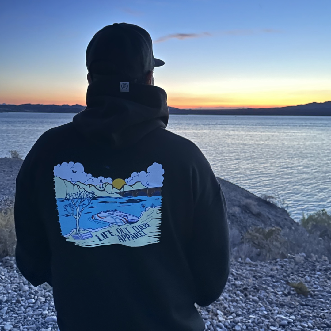 A person wearing the Unisex The Havasu Hoodie in black from Life Out There Apparel enjoys a scenic sunset over Lake Havasu, with rocky shores and distant hills at Windsor Beach against a vibrant sky.