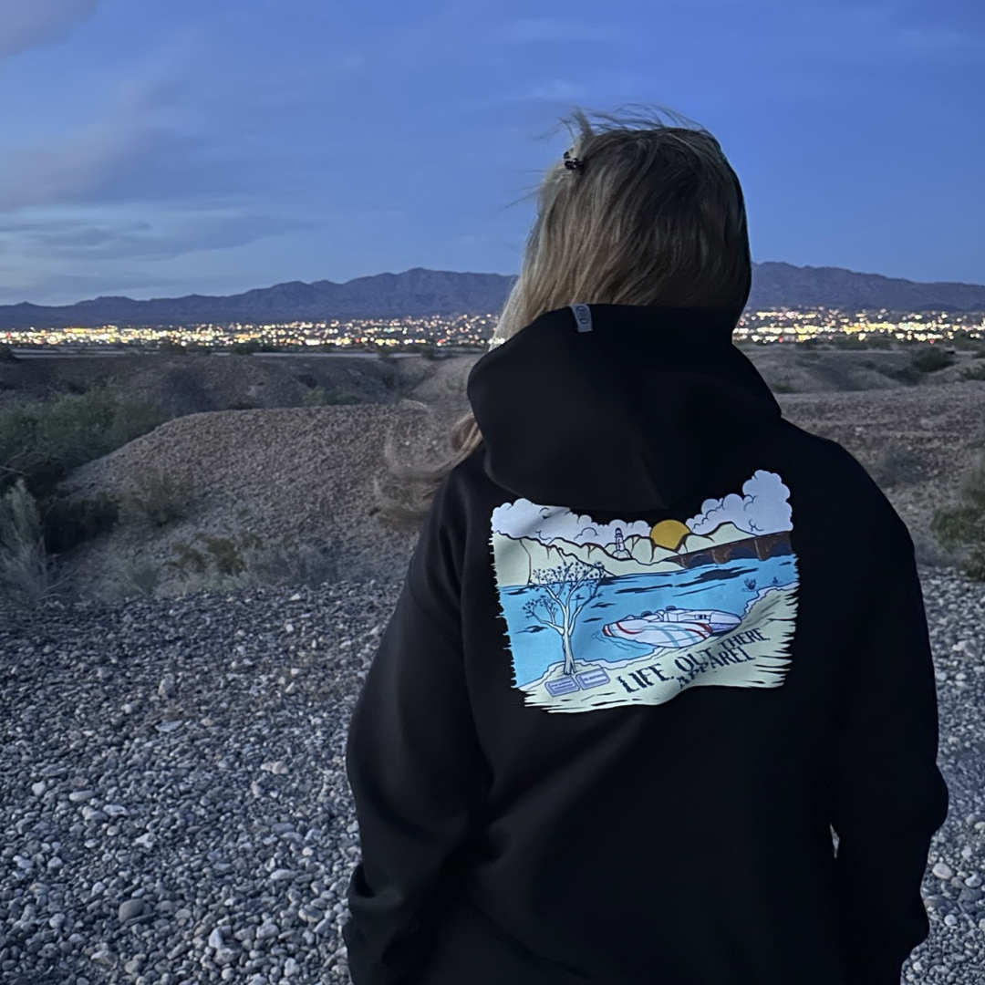 A person dressed in the Unisex The Havasu Hoodie - Black from Life Out There Apparel stands on a rocky hill, gazing at the city skyline of Lake Havasu at dusk. The sky is a gradient of blue, and distant mountains hint at evenings spent near London Bridge or relaxing on Windsor Beach.