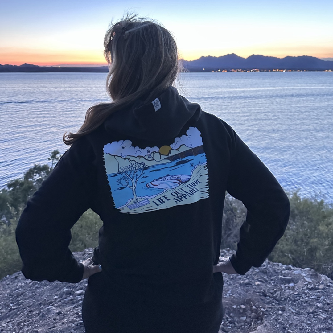 A person stands on a rocky shoreline, capturing the essence of summer as they gaze at the calm sea during sunset. They are wearing Life Out There Apparel's Unisex The Havasu Hoodie in black, featuring a vibrant sunset graphic and the phrase "Life's Good Here," with mountains silhouetted in the background—a nod to carefree days spent wakeboarding at Lake Havasu.