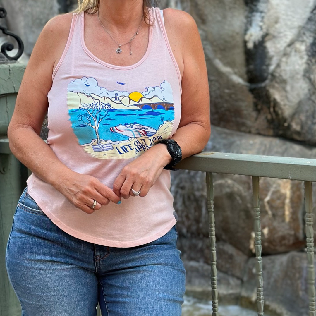 A person, dressed in the Vintage Pink "Havasu Women's Tank Top" by Life Out There Apparel, stands by a railing capturing those summer vibes. They're wearing jeans, a watch, and a necklace. The background showcases rocky textures evocative of Lake Havasu's rugged charm.