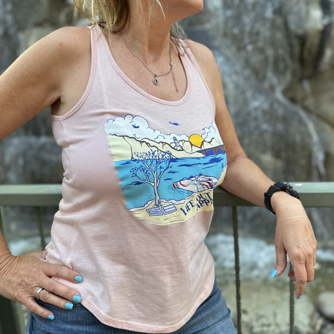 A person wearing Life Out There Apparel's The Havasu Women's Tank Top in vintage pink, adorned with Lake Havasu's sun, clouds, water, and a boat beneath a tree image, leans against a railing while embracing the summer vibes with a waterfall in the background.