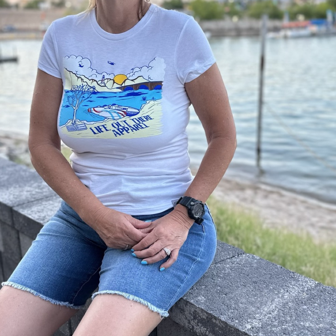A person wearing The Havasu Women's T Shirt in white from Life Out There Apparel, featuring a colorful graphic design, pairs it with denim shorts as they sit on a stone ledge by the calm Lake Havasu. The scene in the background includes trees and a sandy shore beneath the clear sky, perfectly capturing those summer vibes.