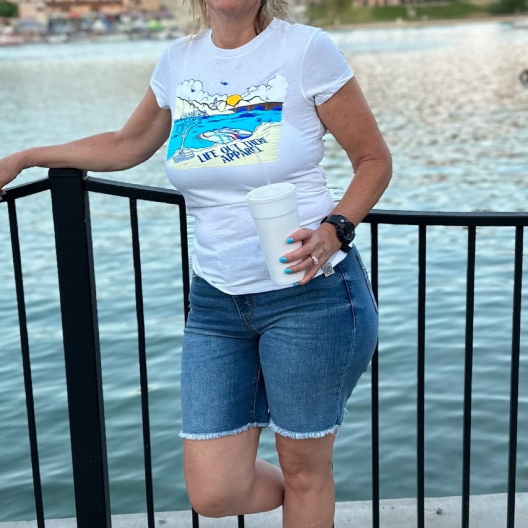 A person dressed in The Havasu Women's T Shirt - White by Life Out There Apparel and denim shorts leans against a railing by Lake Havasu, holding a white cup. In the background, calm water stretches out toward the distant shore of Windsor Beach, accented with buildings.