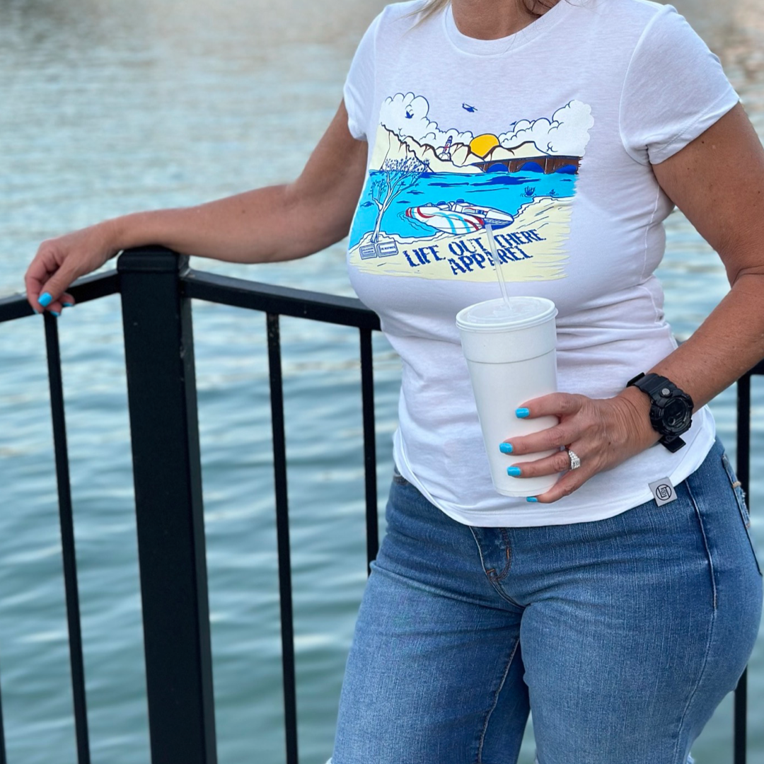 A woman stands by a black railing overlooking the waters of Lake Havasu. Wearing *The Havasu Women's T Shirt - White* from Life Out There Apparel, which features a colorful beach graphic, paired with jeans and holding a large white cup, she exudes summer vibes. Her nails are painted blue under the sunny sky.