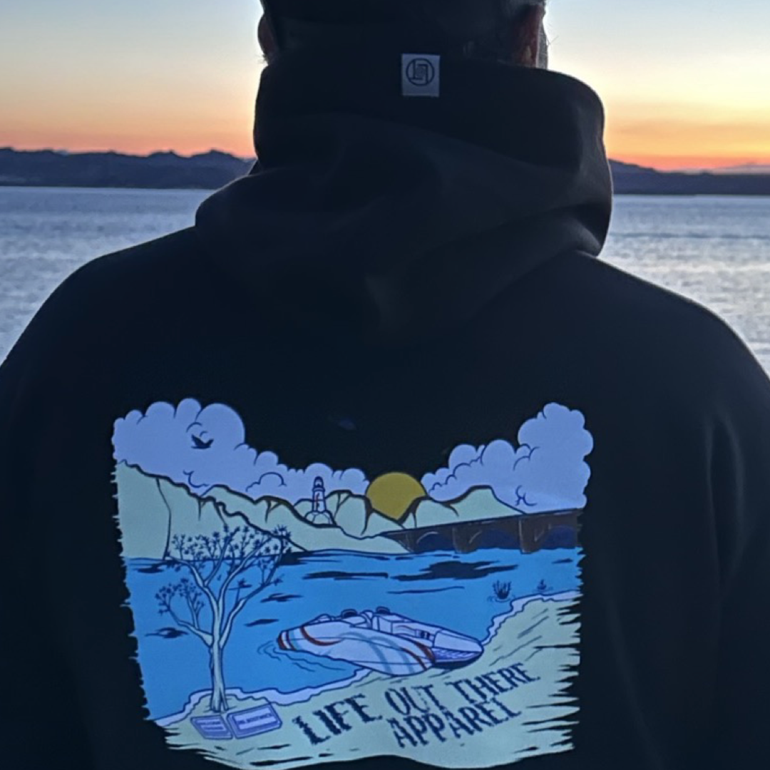 A person dressed in the Unisex The Havasu Hoodie - Black from Life Out There Apparel stands gazing at the sunset over Lake Havasu. The hoodie showcases a vibrant landscape design featuring a tree, mountains, and the brand's name, harmonizing perfectly with the natural beauty and tranquil waters of Windsor Beach.