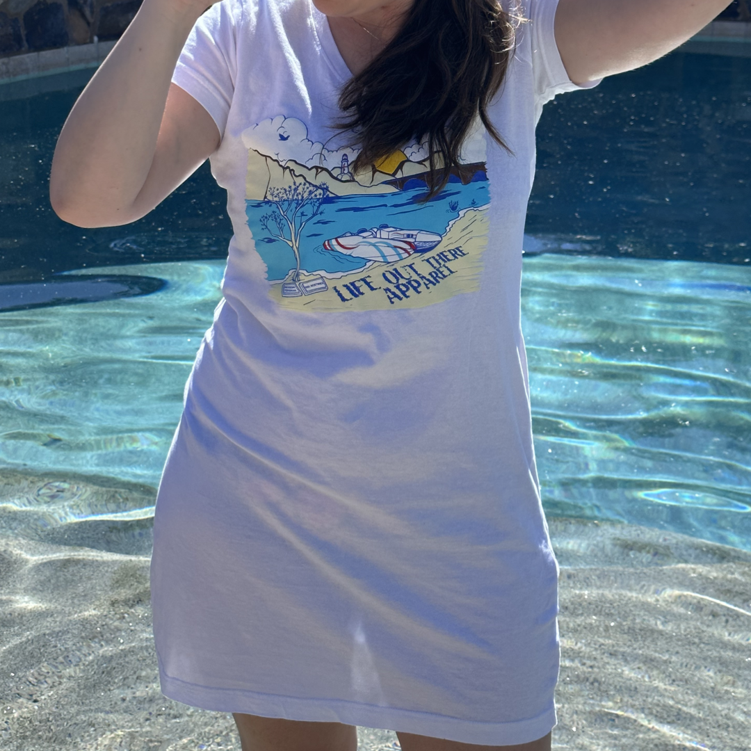 A person stands in a pool, wearing The Havasu Women's Cover Up in white by Life Out There Apparel, featuring a vibrant graphic of Lake Havasu with mountains, trees, and a boat. The sparkling water in the sunlight perfectly captures the summer vibes.