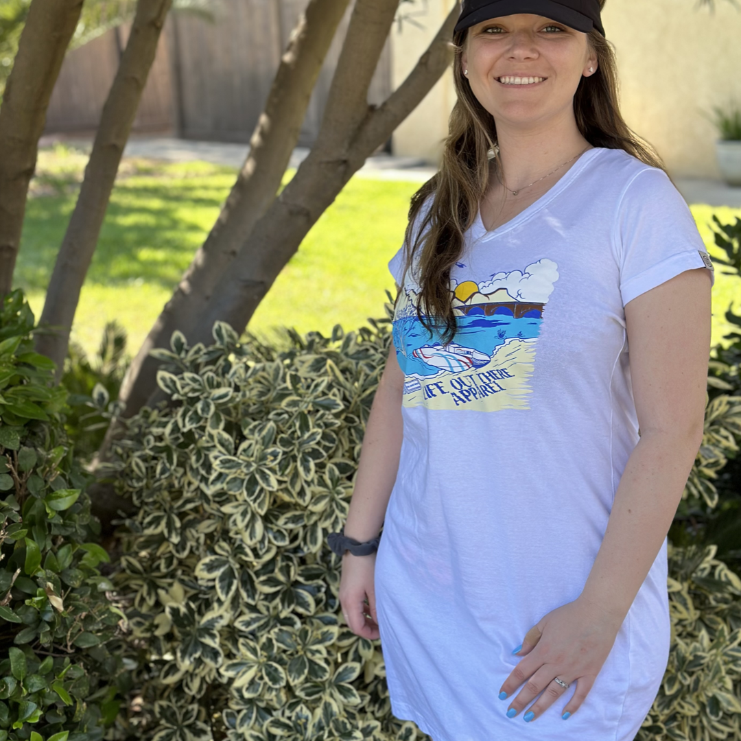 A woman smiles while standing outdoors, embodying summer vibes among lush foliage. She is wearing The Havasu Women's Cover Up in white from Life Out There Apparel, which showcases a vibrant wave and beach scene graphic. A black cap adorns her head, with sunlight creating lively shadows on the grass near the memorial tree behind her.