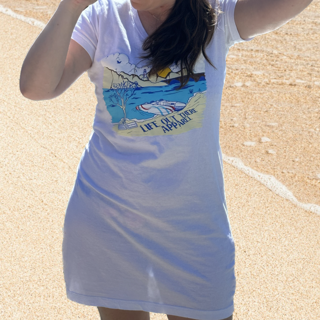 A person standing on a sandy beach by Lake Havasu sports The Havasu Women's Cover Up in white by Life Out There Apparel, showcasing a bright beach scene and the phrase "LIFE OUT THERE ADVENTURE." The summer vibes are unmistakable as waves gently touch the shore in the background.