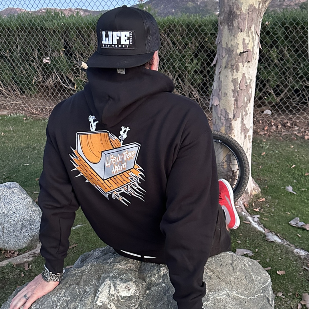 Life Out There Apparel “Fun is Not a Crime” Black Unisex hoodie has a skateboarder and bmx rider having fun on a half pipe skateboard ramp. A man sitting on a rock showing the back of the hoodie.