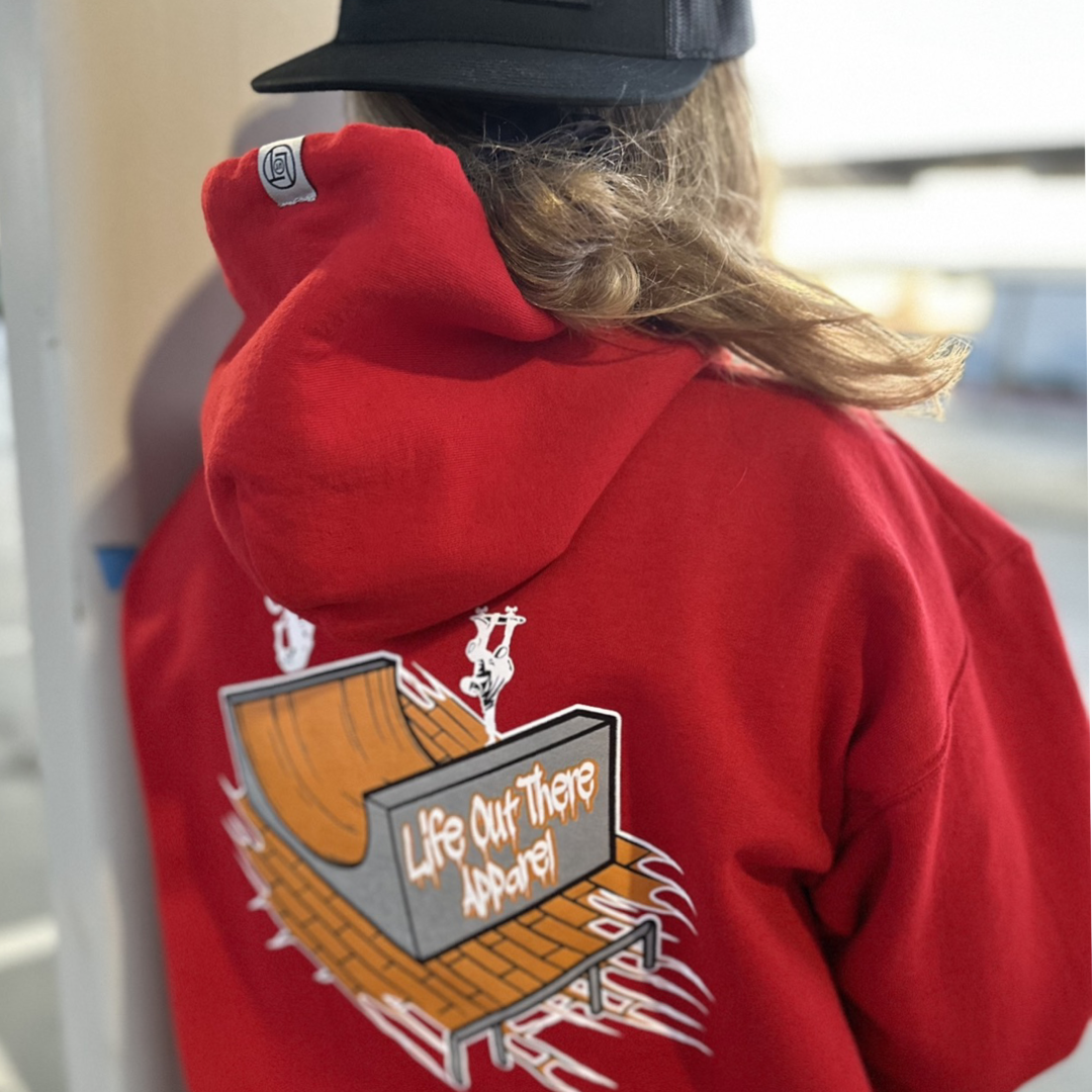 Life Out There Apparel “Fun is Not a Crime” Red Unisex hoodie has a skateboarder and bmx rider having fun on a half pipe skateboard ramp. A woman showing the back design of the hoodie leaning up against a concrete pillar.