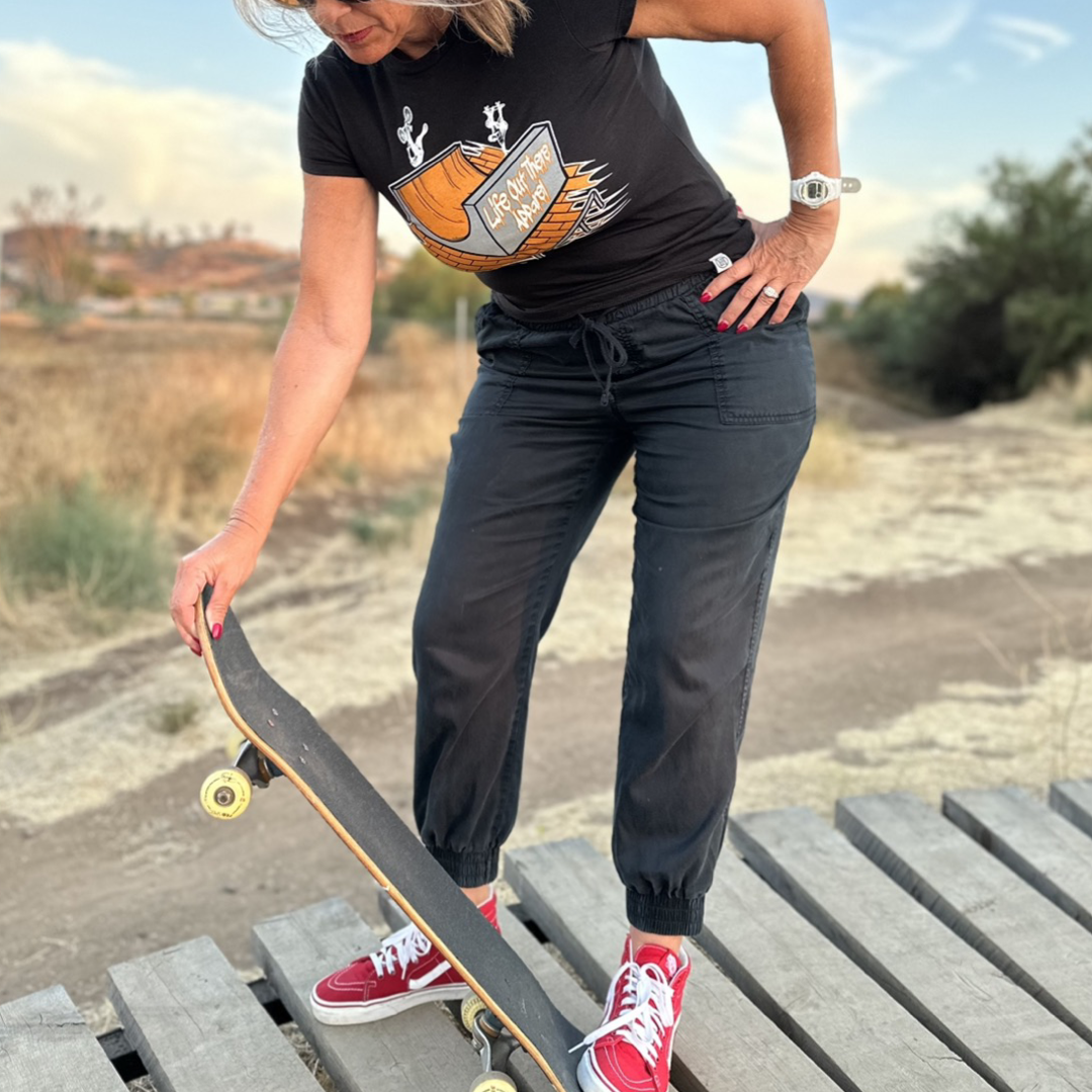 A skateboarder in a Fun Is Not A Crime Women's T Shirt - Black from Life Out There Apparel, paired with black pants and red sneakers, holds a skateboard on a wooden platform. The scene captures the lively essence of the California Amateur Skateboard League against a backdrop of dry landscape, trees, and grass beneath the blue sky.
