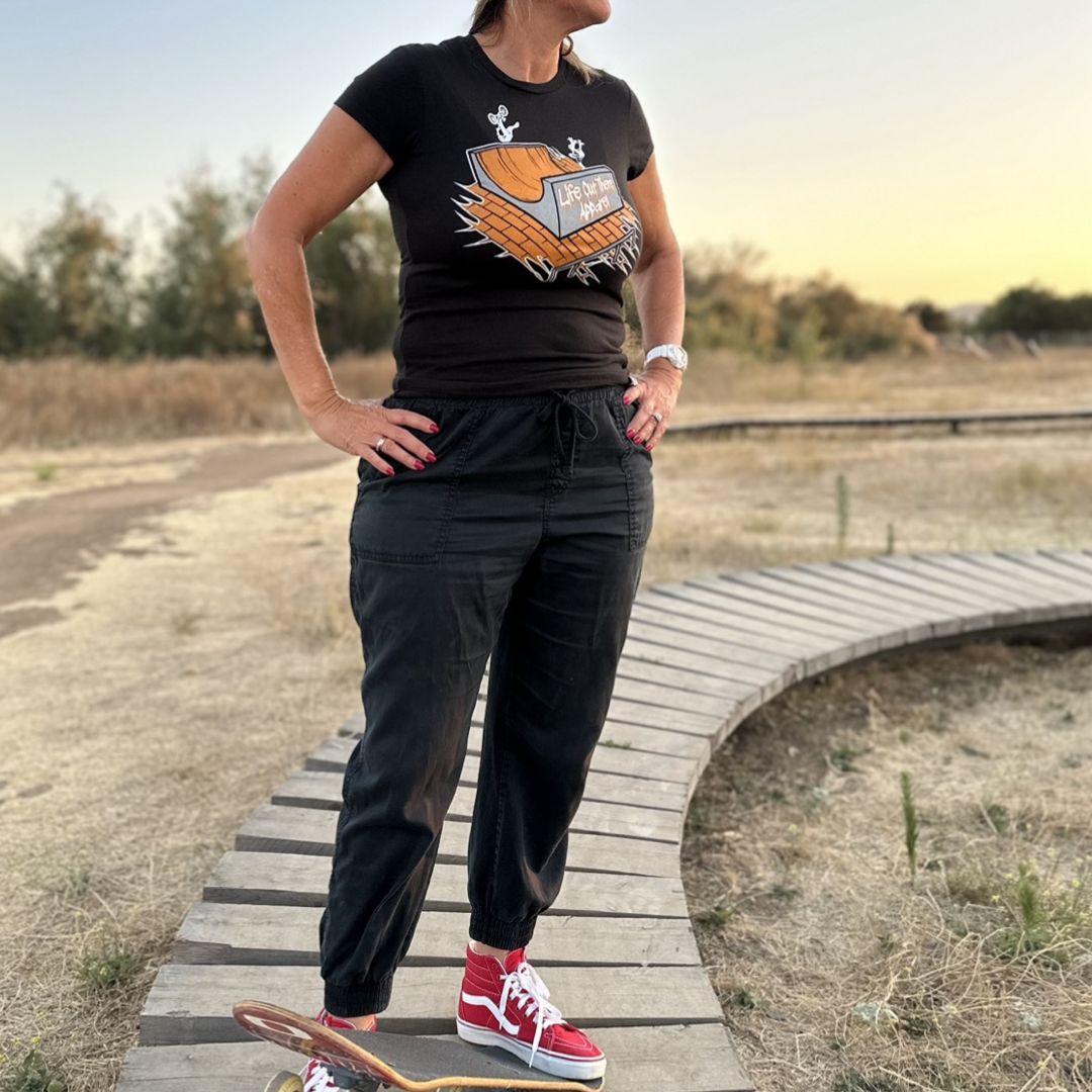 A person stands confidently on a curved wooden path in an outdoor park, sporting the "Fun Is Not A Crime Women's T Shirt - Black" by Life Out There Apparel, paired with black pants and red high-top sneakers. A skateboard rests beside them as BMX bikes zip by in the background against dry grass and trees under the clear sky.
