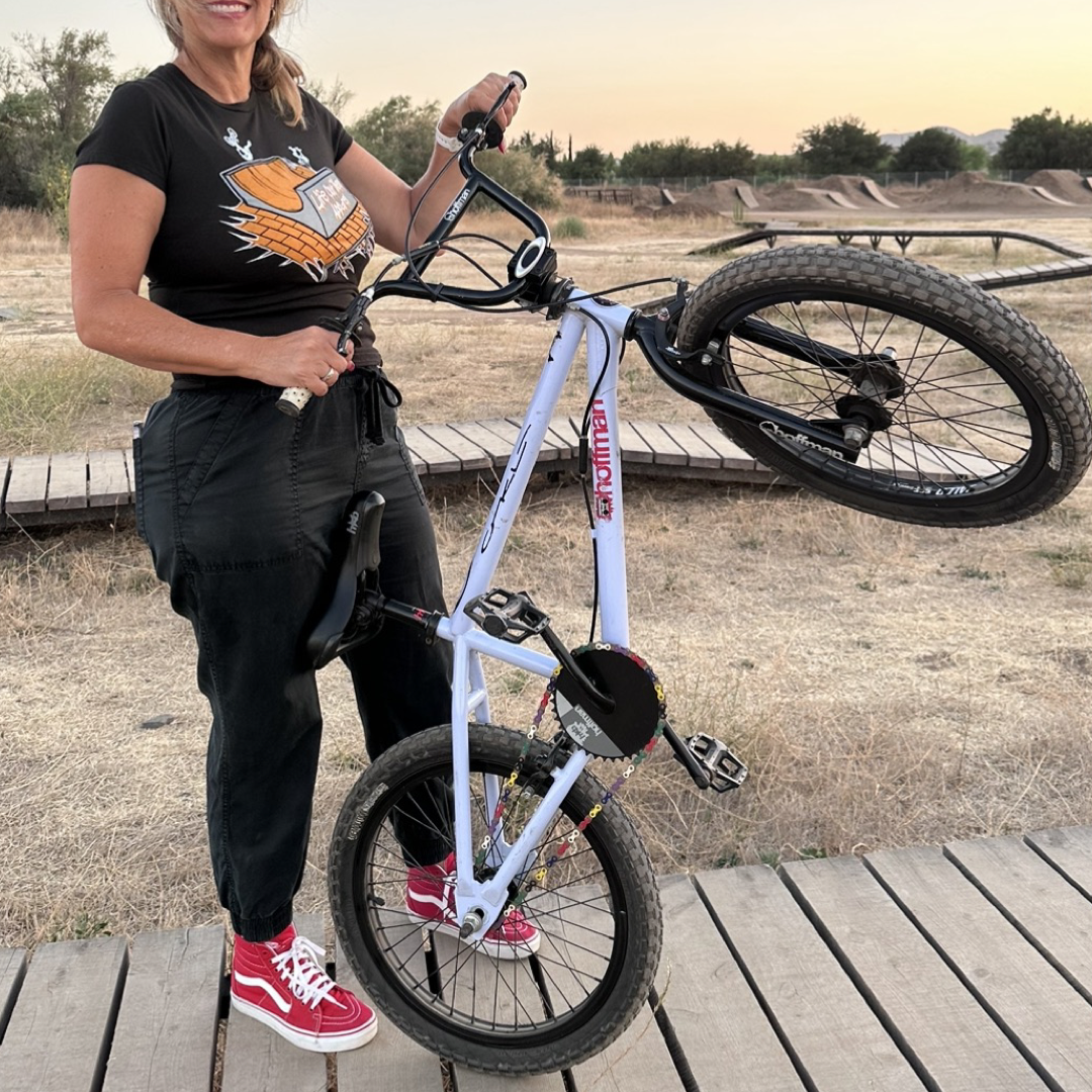 A person stands on a wooden platform holding a BMX bike vertically, preparing for the C.A.S.L. contest. They are wearing Life Out There Apparel's "Fun Is Not A Crime" Women's T-Shirt in black, paired with black pants and red high-top sneakers. Behind them lies a dirt track and grass under a clear sky.