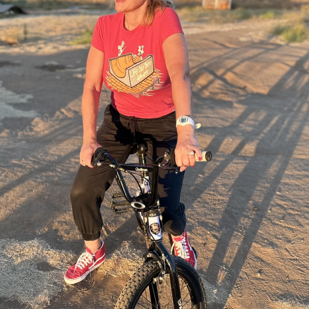 A person dressed in a Fun Is Not A Crime Women's T Shirt by Life Out There Apparel and black pants is sitting on a black bicycle, reminiscent of BMX bikes. The sun casts long shadows on the ground, adding to the vibrant scene. The t-shirt features a colorful design, and the person is poised on a dirt path.