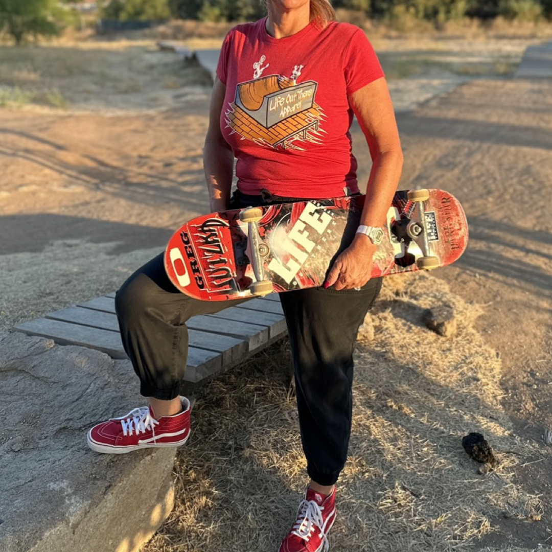 A person wearing the *Fun Is Not A Crime Women's T Shirt* in red from *Life Out There Apparel* and black pants holds a skateboard, standing on a dirt path with one foot resting on a stone. The sun casts long shadows, suggesting an ideal setting for the California Amateur Skateboard League, as trees provide a picturesque backdrop.