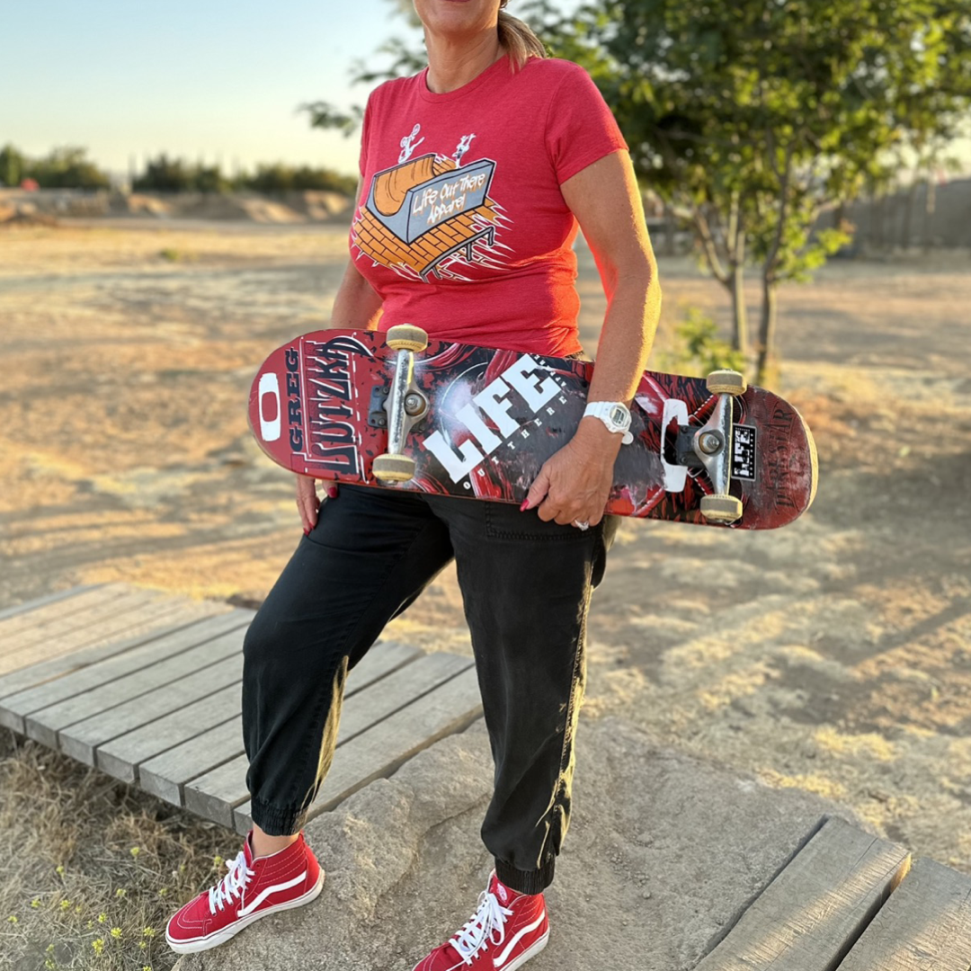 A person wearing a Fun Is Not A Crime Women's T Shirt in red from Life Out There Apparel and black pants stands outdoors holding a skateboard with a LIFE design. They're sporting red sneakers on wooden planks, set against a dry, grassy California landscape dotted with trees—a perfect backdrop for any California Amateur Skateboard League event.
