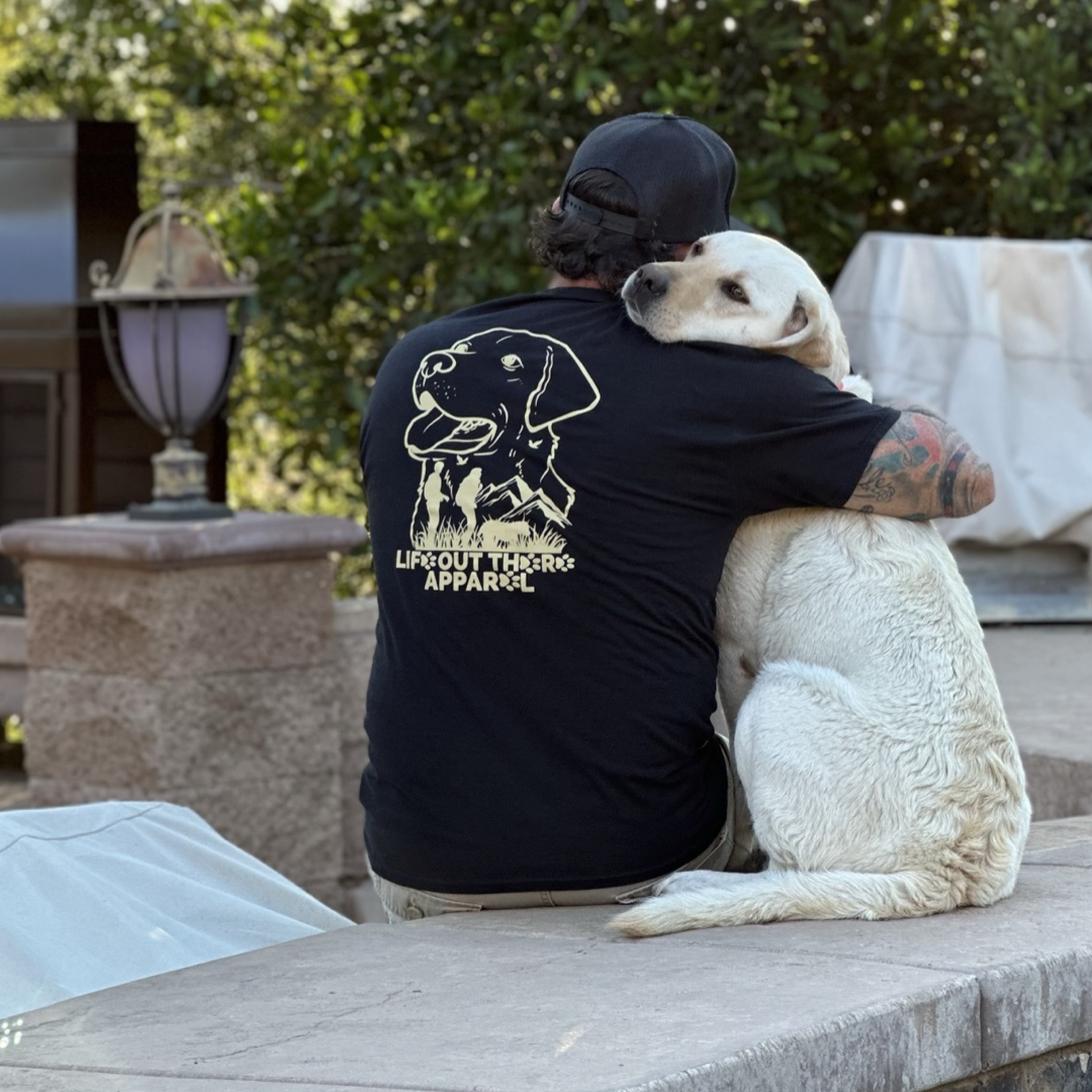 A man with a tattooed arm wearing the Life Out There Apparel Unisex Best Friend Tee - Black, featuring a dog illustration, sits on a ledge. He embraces a large white Labrador retriever, symbolizing unconditional love as they both gaze into the distance. Behind them, a patio adorned with greenery and illuminated by a lamp provides the backdrop.