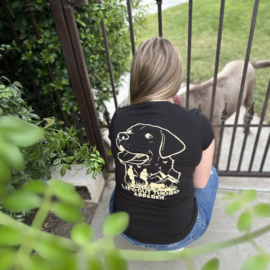 Dressed in a Best Friend Women's T-Shirt - Black by Life Out There Apparel, someone sits peacefully by a garden gate. On the other side, their loyal canine companion waits patiently. Surrounded by green plants, the scene beautifully captures the essence of unconditional love shared in this tranquil moment.