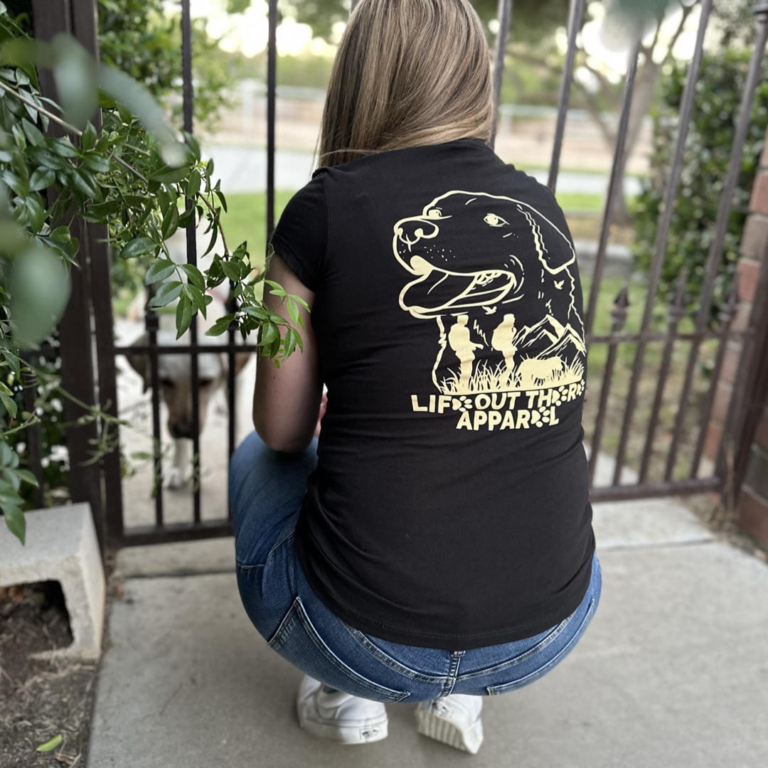 Next to a gate, a person crouches wearing the Life Out There Apparel Best Friend Women's T-Shirt in black, which showcases a dog graphic. Beyond the gate, their energetic canine companion looks on. The vibrant greenery and brick pillar further enhance this outdoor scene, embodying the spirit of unconditional love.