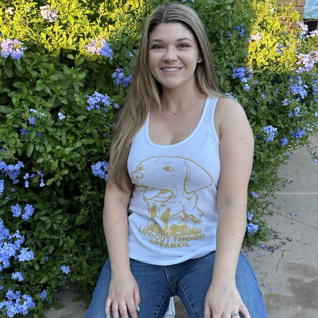 A person with long hair sits in front of a bush with blue flowers, wearing the Best Friend Women's Tank Top by Life Out There Apparel. In this sunny garden setting, they embrace their best friend, celebrating a love for dogs and nature.