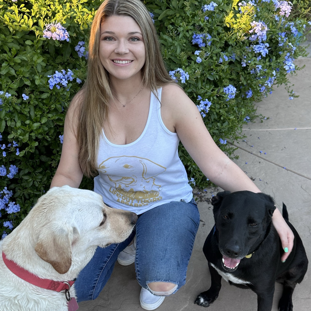 A woman kneels on a patio, her face beaming with affection as she wraps her arm around her black dog, her best friend. A yellow dog faces them warmly. She is wearing the "Best Friend Women's Tank Top" in white by Life Out There Apparel, paired with blue jeans and surrounded by the vibrant embrace of purple-flowered bushes.