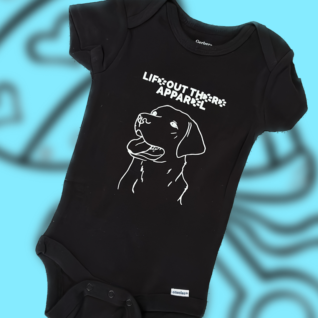 The Unisex Best Friend Baby Onesies in black, made from 100% cotton, features a white line drawing of a dog's face. The text "Life Out There Apparel" is displayed above, and the background includes a blurred blue version of the dog drawing, celebrating your little one's best friend in style.
