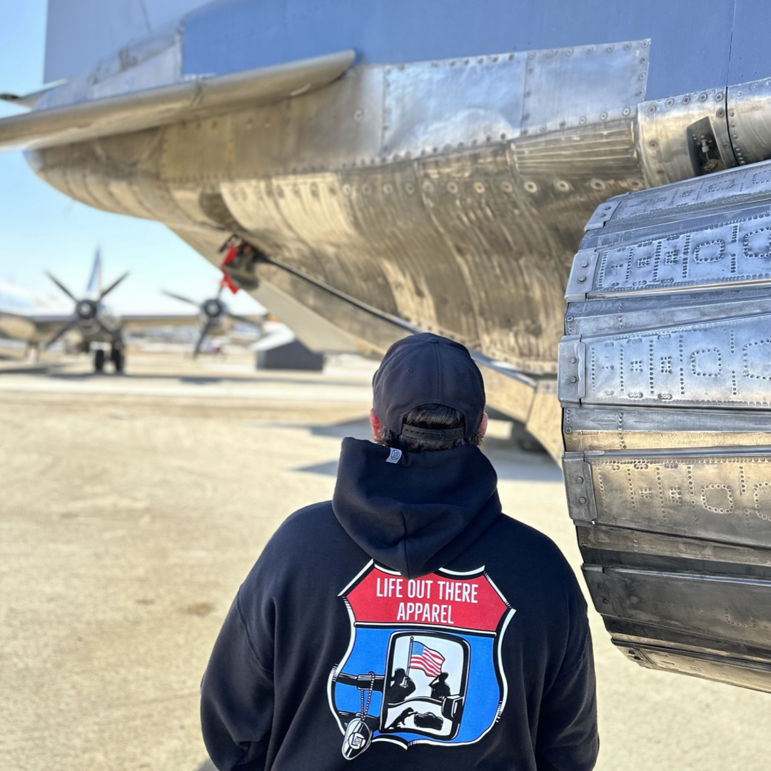 Life Out There Apparel “Leave It Behind” Unisex “Zip Up” hoodie has a truck side mirror with the reflection of a male, female & K9 soldiers saluting the American flag - bringing awareness to P. T. S. D. (post traumatic stress disorder). This is a photo of a man wearing the hoodie showing the back design, standing next to a military airplane.