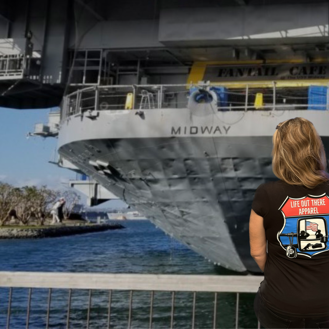 A woman in a Leave It Behind Women's T-Shirt by Life Out There Apparel, featuring a beach scene design on a black fabric, stands by the railing as she gazes at the USS Midway, an aircraft carrier docked at the harbor. On this Veterans Day, her reflection is framed by the clear sky and trees, honoring those who served.