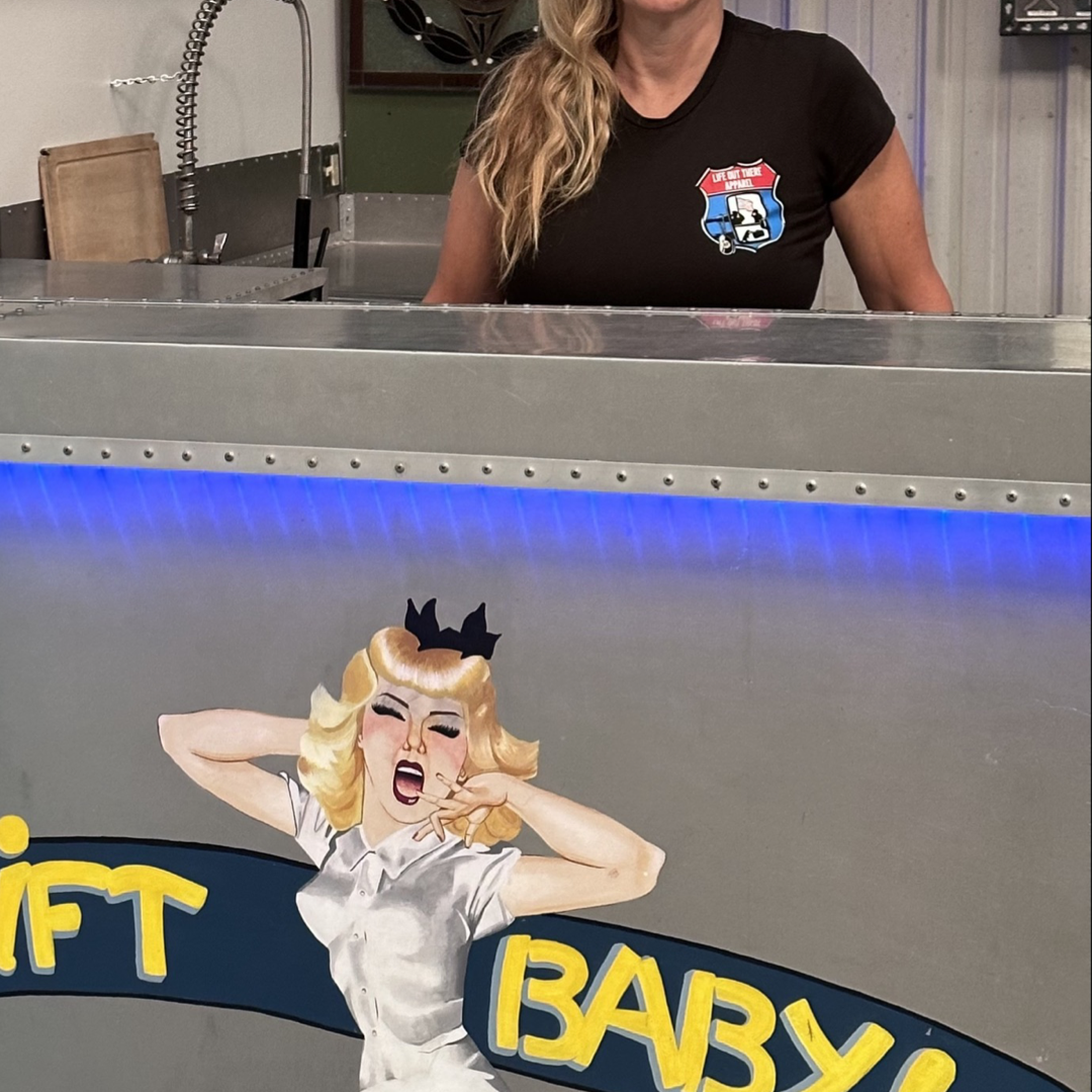 A person with long blonde hair stands behind a metallic counter, where a painted image of a woman in a white dress is captioned with the words "Swift Baby!" In the background, industrial elements like metal and equipment subtly pay homage to veterans. They are wearing the Life Out There Apparel's "Leave It Behind Women's T Shirt" in black.