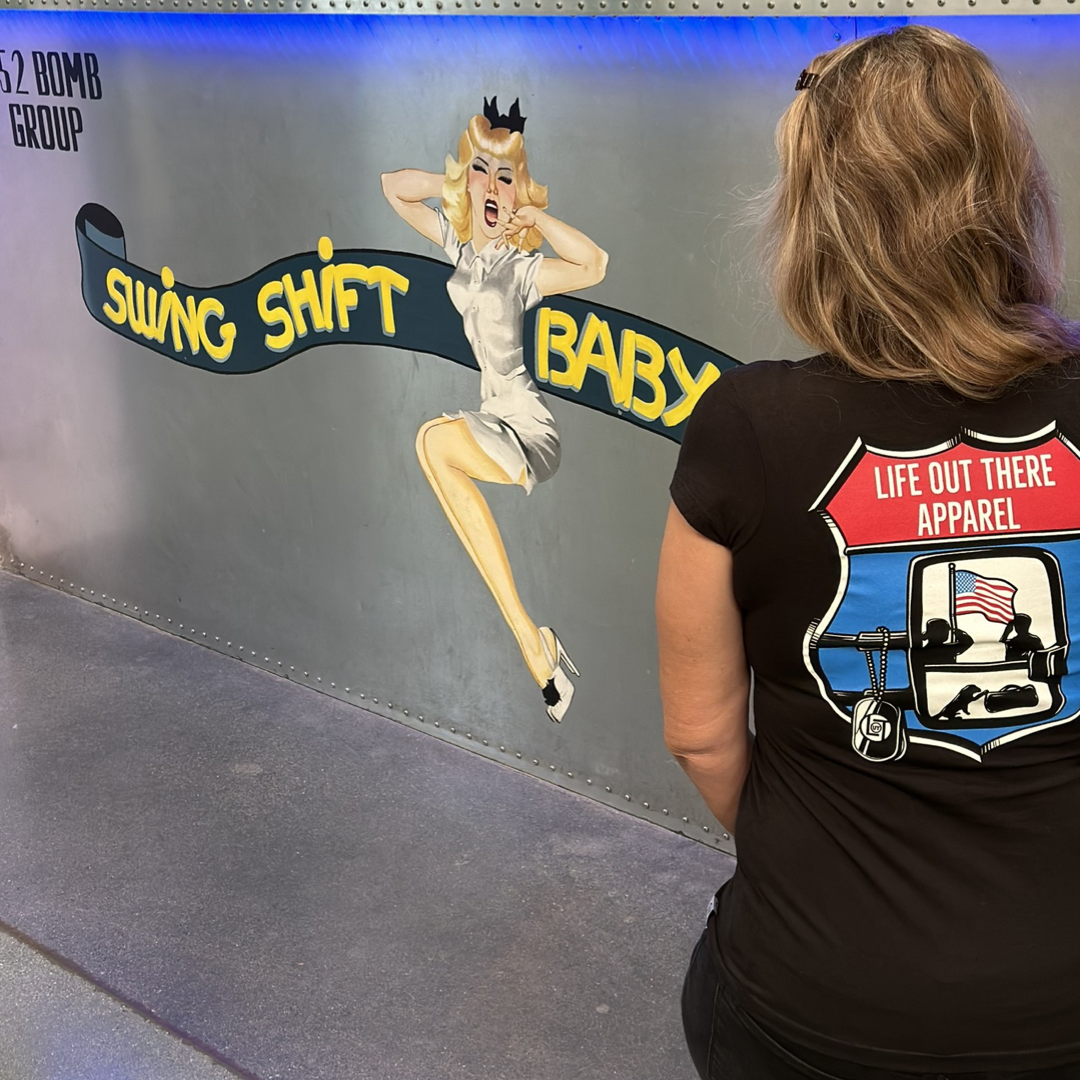A woman stands facing a mural of a vintage pin-up girl with the words "Swing Shift Baby" beside her. In honor of Veterans Day, she wears the "Leave It Behind" Women's T Shirt in black by Life Out There Apparel, featuring a golf cart adorned with an American flag, paying tribute to veterans everywhere.