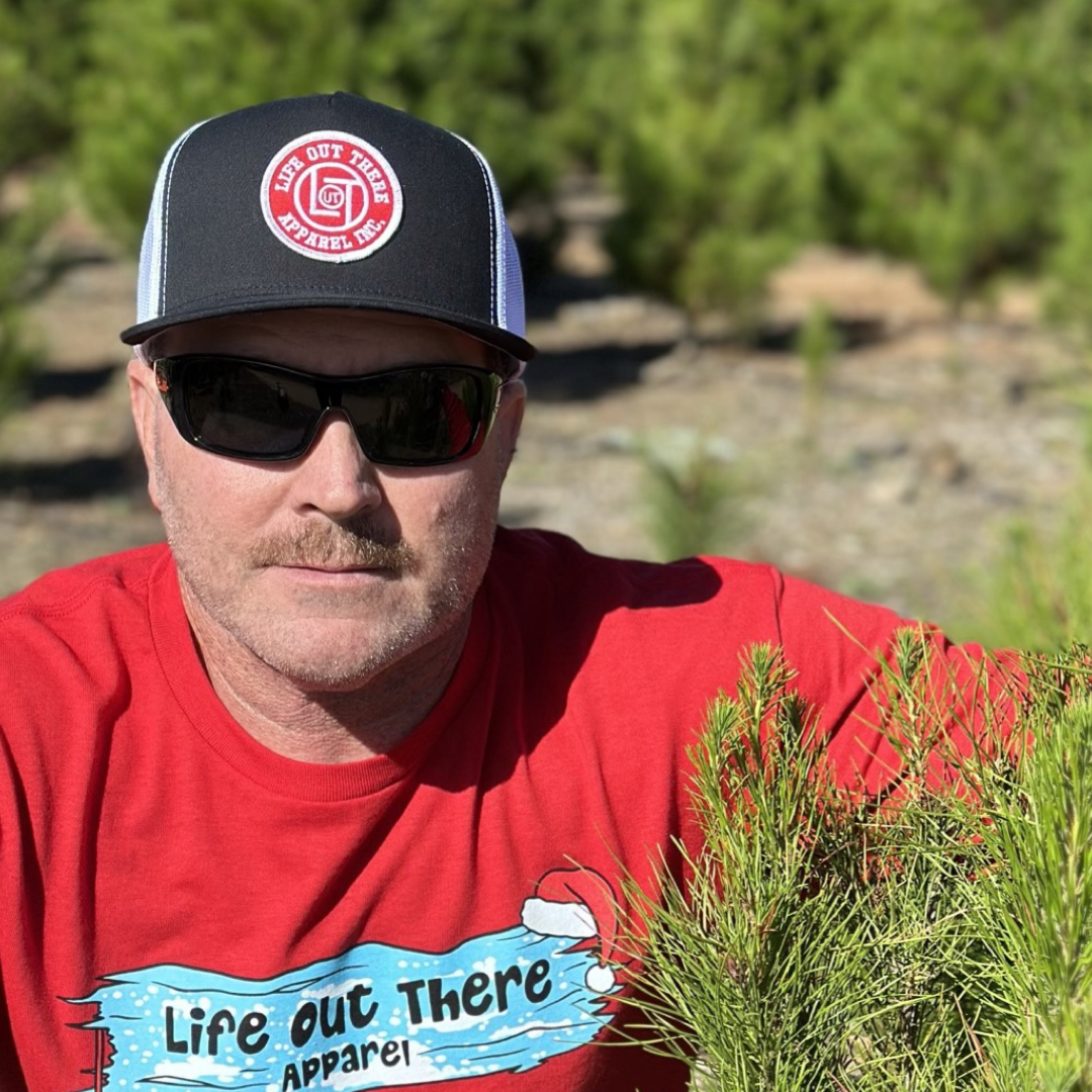 A person wearing a red shirt, black cap, and sunglasses stands outdoors in a sunny, wooded area. The LOT Snapback "Flat Bill" Trucker Hat by Life Out There Apparel adds style as they pose near vibrant green foliage, looking directly at the camera.