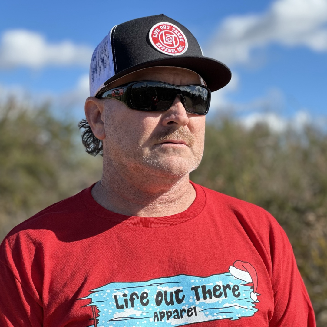 A man wearing a red T-shirt from Life Out There Apparel, complemented by a LOT Snapback "Flat Bill" Trucker Hat in black and white and sunglasses, stands outdoors against a backdrop of bushes and blue skies with clouds.