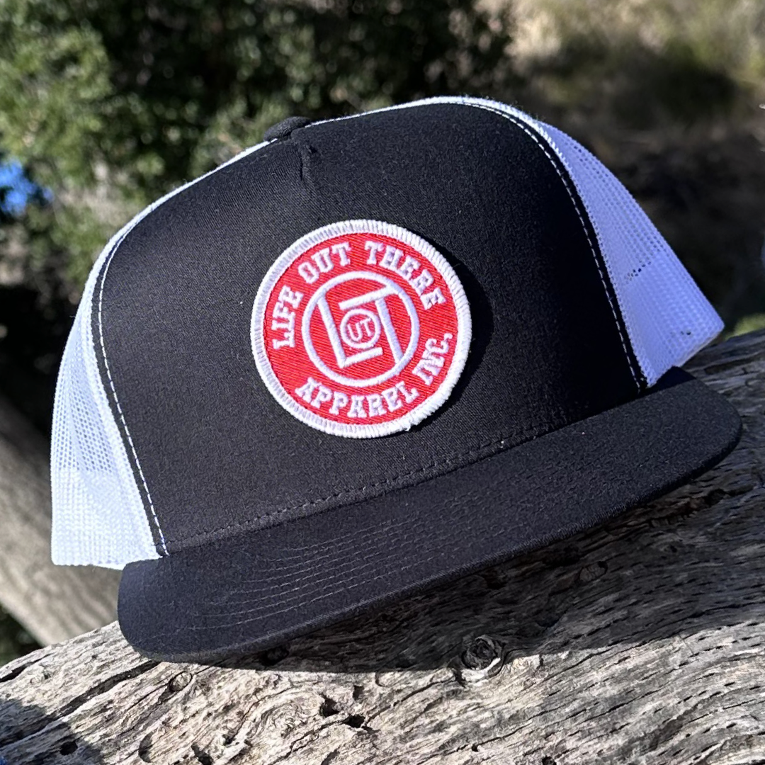 A LOT Snapback "Flat Bill" Trucker Hat - BLACK/WHITE from Life Out There Apparel, featuring a red circular logo patch, is placed on a wooden surface. The cap's design is enhanced by the blurred greenery and sunlight in the background.