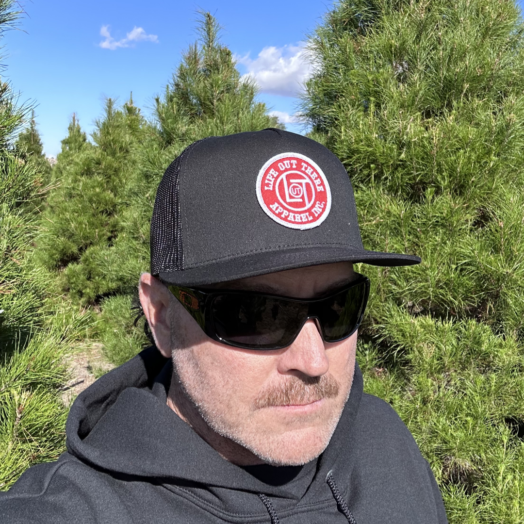 A person wearing a black LOT Snapback "Flat Bill" Trucker Hat from Life Out There Apparel and sunglasses stands among evergreen trees. They're dressed in a black hoodie, and the cap features a red circular logo with the text Life Out There Apparel Co. The sky is clear and blue above.
