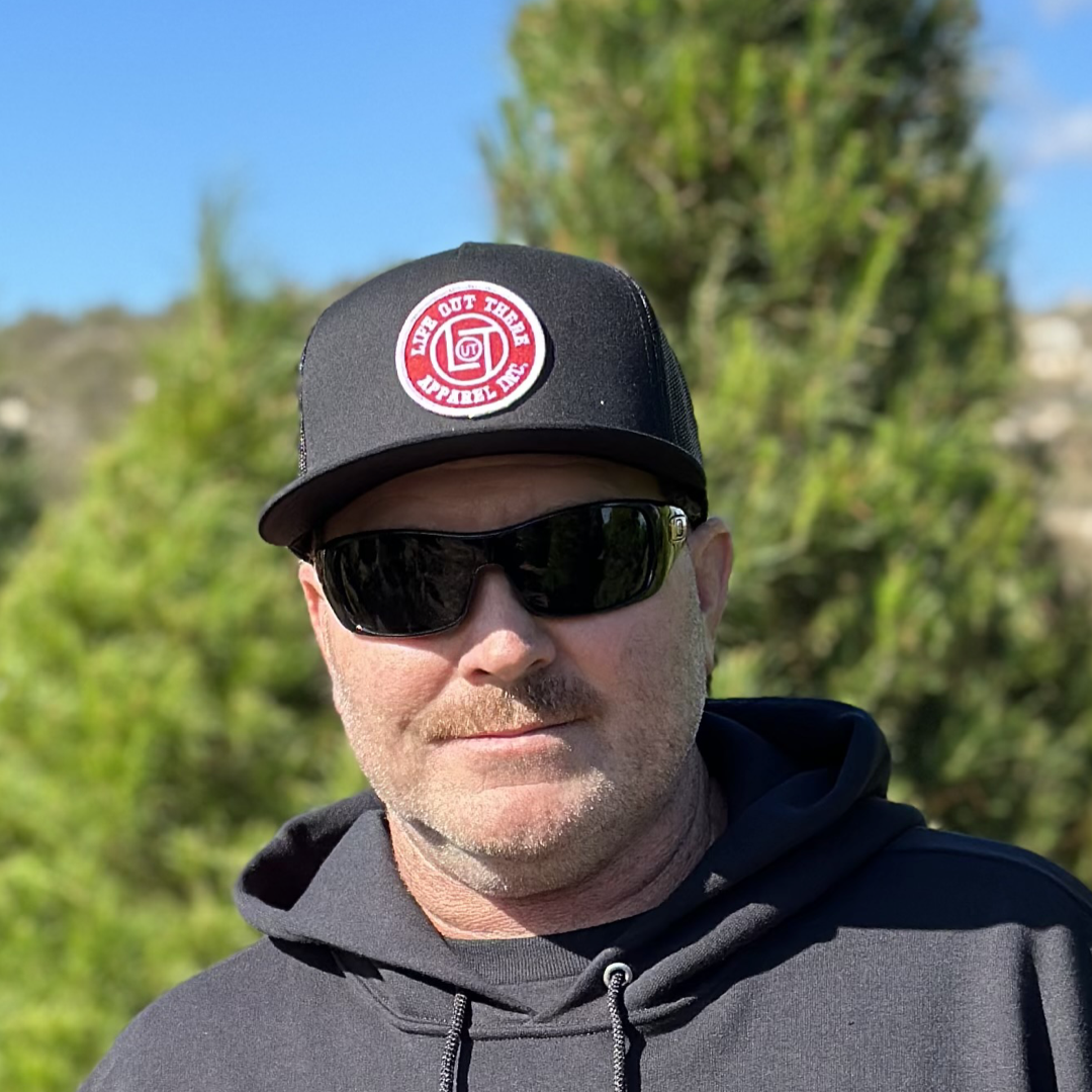 A man wearing the LOT Snapback "Flat Bill" Trucker Hat in black by Life Out There Apparel, sunglasses, and a dark hoodie stands outdoors. Greenery and a clear blue sky are in the background.