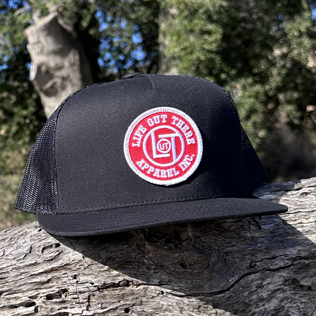 A LOT Snapback "Flat Bill" Trucker Hat in black from Life Out There Apparel, featuring a red and white patch with the brand's name, is placed on a textured piece of wood. The blurred greenery and a tree trunk in the background enhance the rustic charm of the scene.