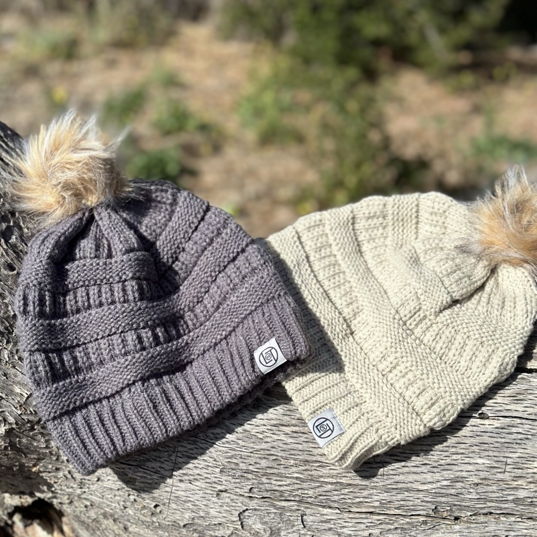 Two Women’s Pom Beanie 2.0 from Life Out There Apparel rest on a weathered wooden surface, each adorned with faux fur pom-poms. One beanie is a charcoal grey color, while the other is beige, and both display small brand labels on the brim against a blurred outdoor backdrop with greenery.