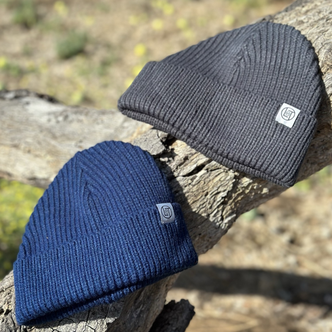 A navy men's knit beanie from Life Out There Apparel rests on a textured tree branch alongside another in charcoal. Both hats boast a small logo patch. The softly blurred background enhances the intricate details of each beanie, allowing them to stand out beautifully.